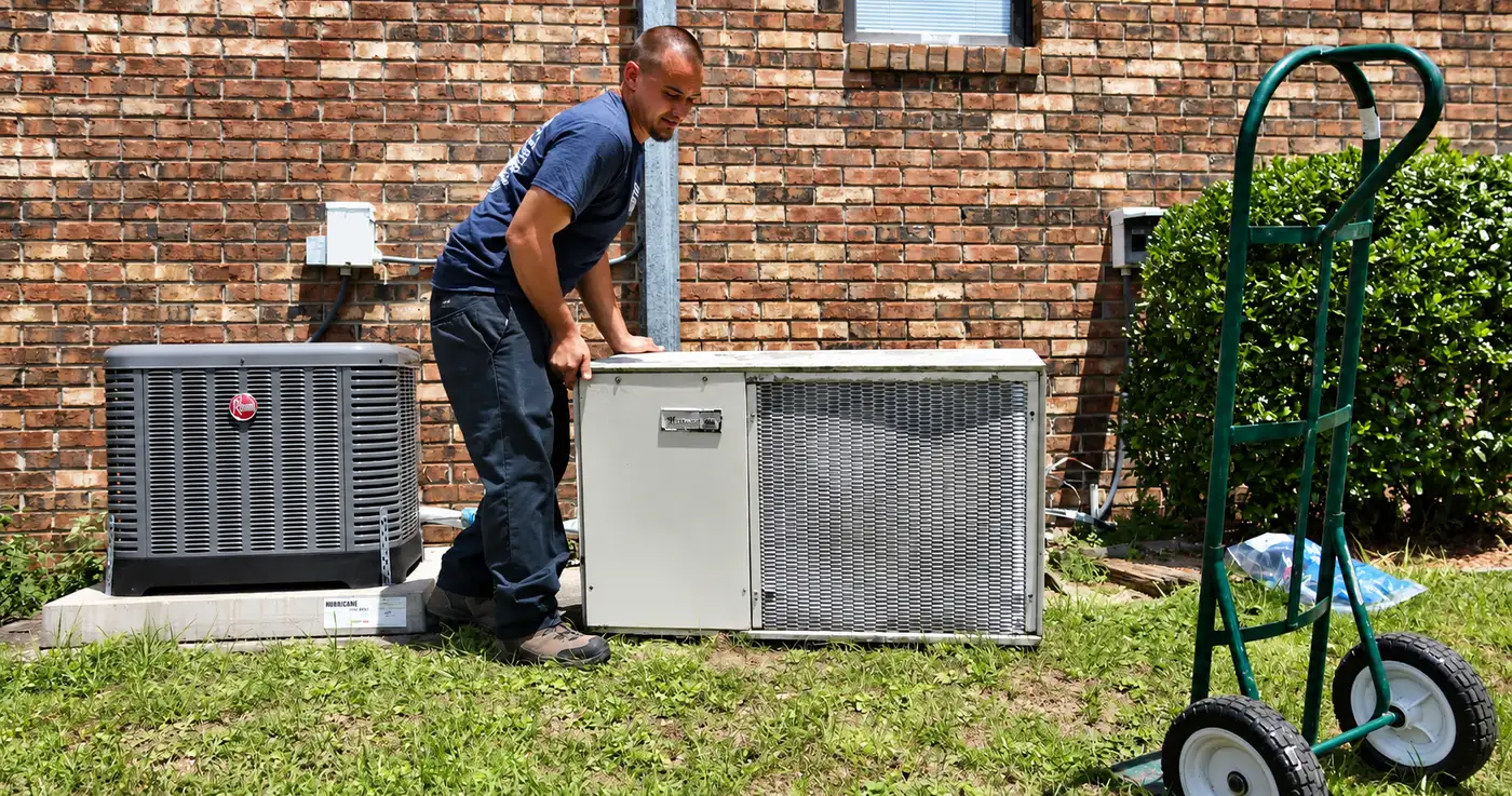 An R.J. Kielty technician moving a replacement air conditioning unit into place beside a home