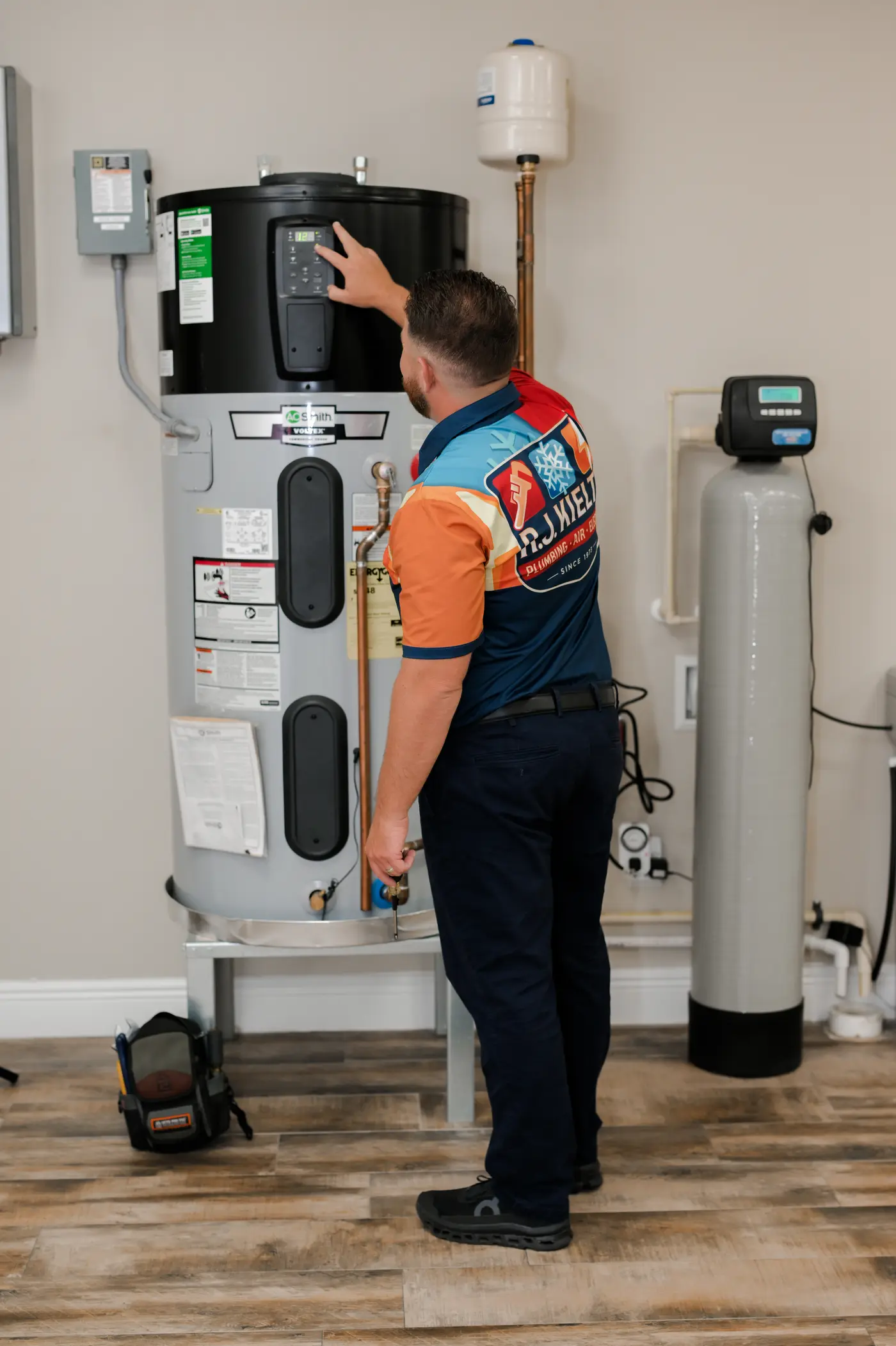 A technician servicing a whole-home water softener beside a water heater in a utility room