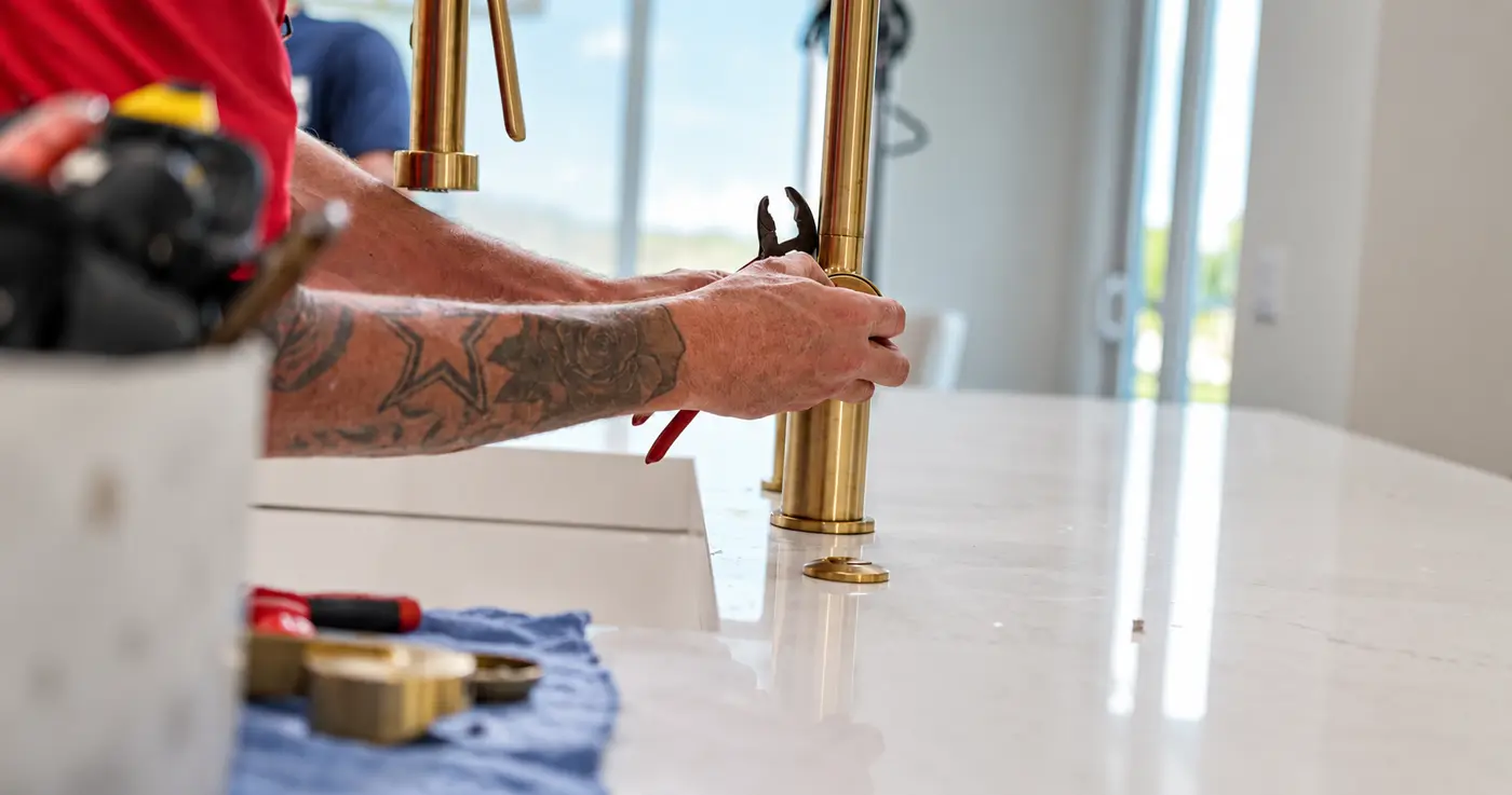 A plumbing technician installing and tightening a brass kitchen faucet at the countertop